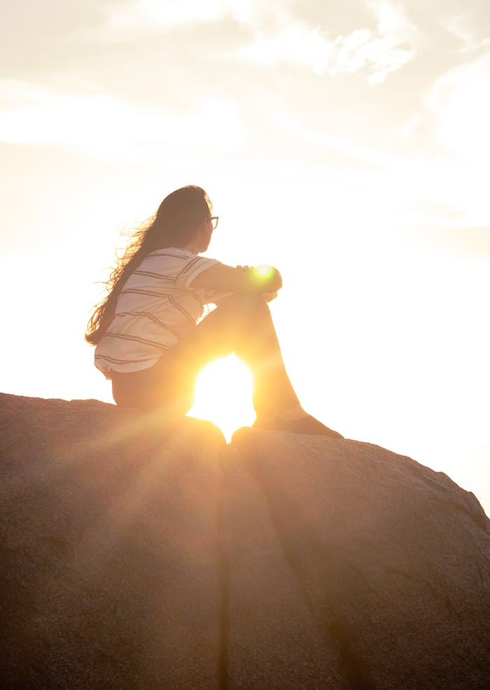 A woman with long hair sitting on a rock, silhouetted against a vibrant sunset.