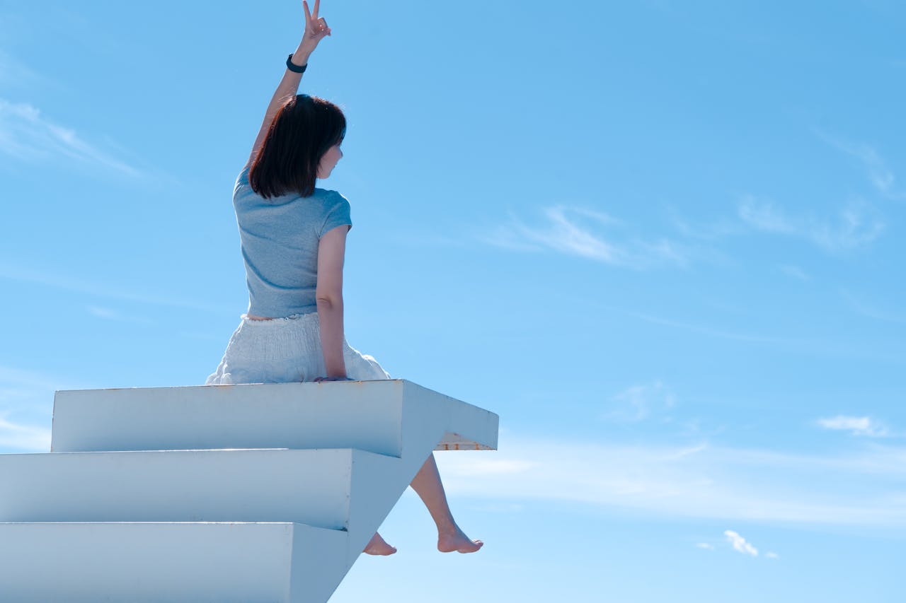 A woman sits on white stairs with blue sky background, reaching upward, embracing freedom.